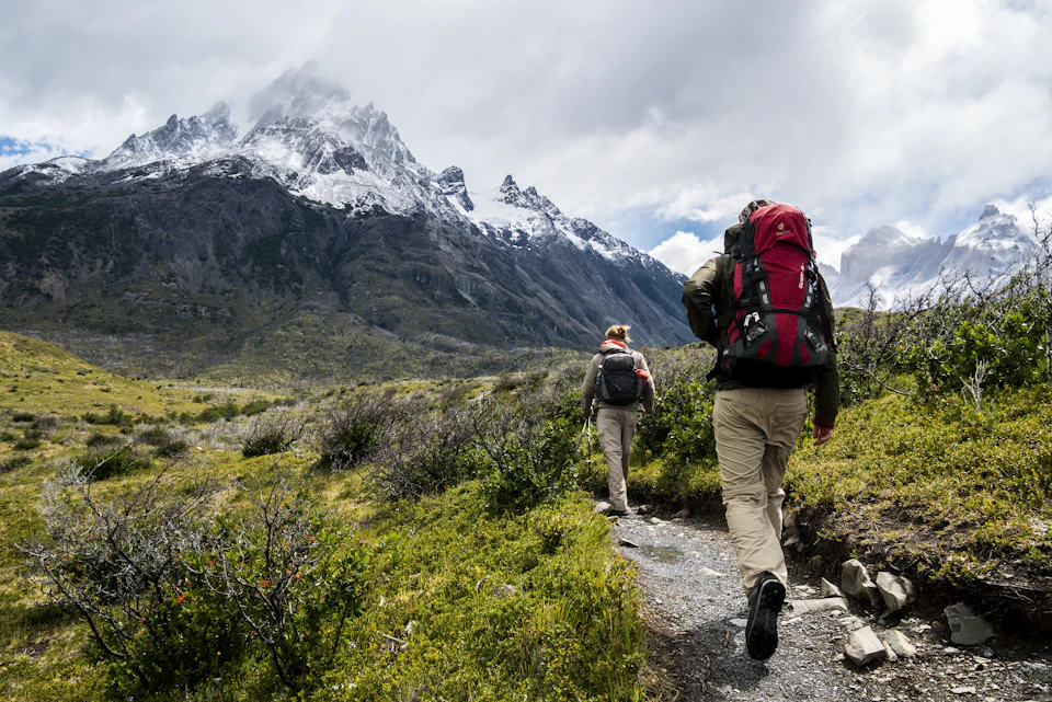 Two hikers with backpacks walking along a trail toward snow-capped mountains